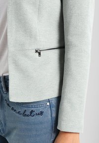 Veste texturée vert clair avec une poche zippée en détail, associée à un jean bleu avec un texte brodé sur la poche.