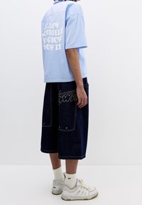 Camicia azzurro chiaro a maniche corte con testo bianco sul retro; pantaloni corti in denim scuro con tasche; sneakers bianche con accenti grigi.