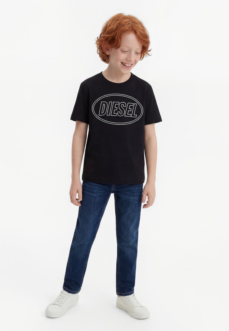 Χαμογελαστό αγόρι με κόκκινα μαλλιά, φορώντας μαύρο μπλουζάκι με το λογότυπο Diesel, μπλε τζιν και λευκά αθλητικά παπούτσια, στέκεται μπροστά σε λευκό φόντο.