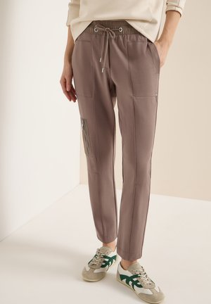 Persona indossa pantaloni jogger taupe con tasca laterale con cerniera e vita con coulisse, abbinati a sneakers bianche e verdi.