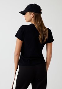 Sort bomulds T-shirt med korte ærmer, udstyret med rund hals. Bæret sammen med sorte bukser, håret falder lige ned over skuldrene, og en sort kasket.