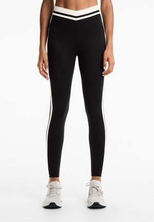 Black leggings with white side stripes, high waistband, fitted design, and smooth texture. Paired with light-colored athletic shoes.