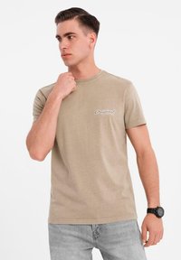 Khaki tričko s krátkými rukávy a kulatým výstřihem s potiskem "Original" na levé straně hrudi. Model má na sobě světle šedé džíny a černé hodinky.