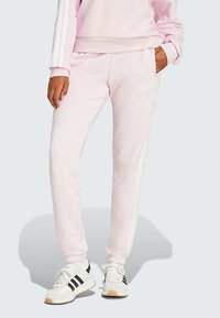 Svijetlo ružičaste trenirke od mekanog materijala, s tri bijele bočne pruge i bočnim džepovima, u kombinaciji s bijelim tenisicama s crnim naglascima.