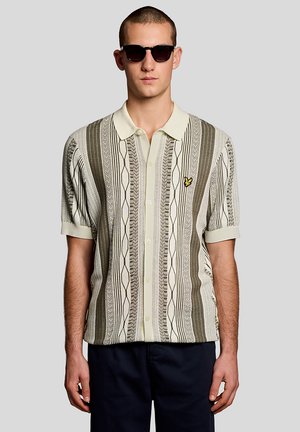 Mann trägt gemustertes, kurzärmeliges Hemd mit Knopfleiste und Kragen, Sonnenbrille und dunkle Hose vor hellem, einfarbigem Hintergrund.