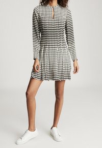 Schwarz-weiß gemustertes Kleid mit langen Ärmeln, einem Ausschnitt im Schlüssellochdesign und einem ausgestellten Rock, kombiniert mit weißen Sneakers.