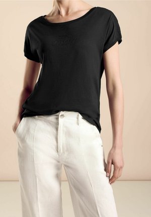 Kvinde iført en sort T-shirt med korte ærmer og subtil tekst samt hvide bukser med høj talje, stående med den ene hånd i lommen mod en beige baggrund.