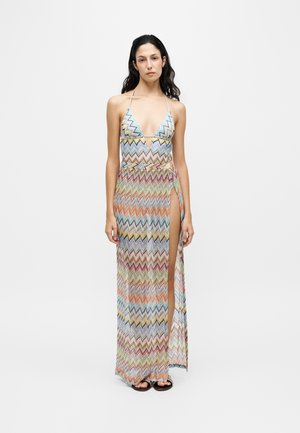 Femme portant une robe longue dos nu à motif zigzag multicolore avec une fente haute sur la cuisse, debout devant un fond uni.