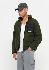 Groene fleece jack met een ritssluiting aan de voorkant, zwarte borstzak en contrasterende stiksels. Gestyled met een witte tanktop en lichtgrijze cargobroek.