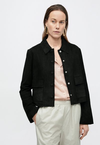 Μαύρο σουέντ cropped μπουφάν με γιακά, ασημένια κουμπώματα τύπου snap και δύο μπροστινές τσέπες, φορεμένο πάνω από ροζ ανοιχτό πουκάμισο με κουμπιά.