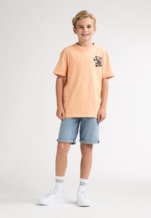 Tricou din bumbac de culoarea piersicii, cu un detaliu grafic pe buzunar, asortat cu pantaloni scurți din denim cu tiv suflecat. Tenisi albi și șosete cu dungi până la gleznă completează ținuta.