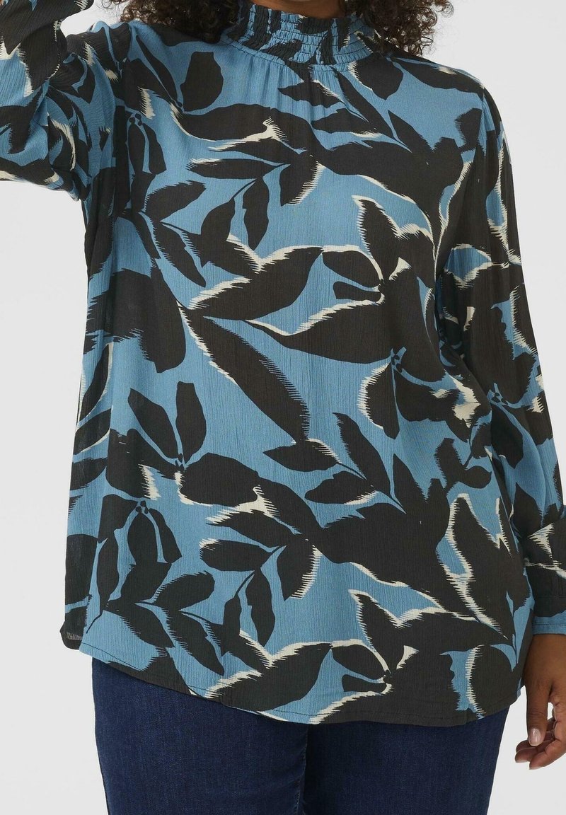 Personne portant un chemisier bleu avec un motif de feuilles abstraites noires et blanches et un jean bleu foncé, montrée des épaules aux hanches sur un fond uni.