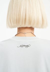 Gray short-sleeve shirt featuring a brown embroidered "Ed Hardy" logo on the back neckline, worn with a silver chain accessory.