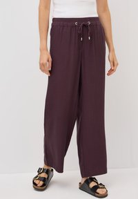 Bordeaux brede broek met een elastische tailleband met trekkoord, voorzien van zilveren oogjes en een lichte textuurstof. Gecombineerd met zwarte sandalen.
