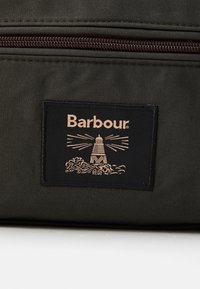 Tecido verde escuro com um emblema retangular preto da Barbour, apresentando um farol e ondas, abaixo de um zíper castanho fechado.