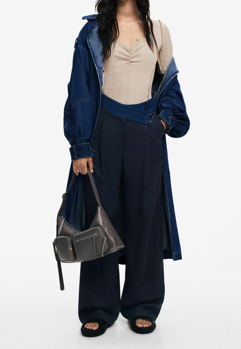 Femme portant un haut beige froncé, un pantalon oversize bleu foncé, une longue veste en jean, des chaussures noires à bout ouvert, et tenant un sac à main gris.