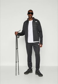 Muškarac u crnom North Face trenirkom i planinarskim cipelama drži planinarske štape, nosi velike crne naočale, stoji na jednoličnoj pozadini.