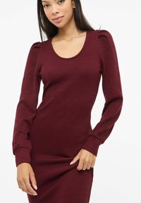 Vestido burdeos de canalé con mangas largas, escote redondo y detalles fruncidos en los hombros; textura suave, silueta ajustada y largo a la altura de la rodilla.