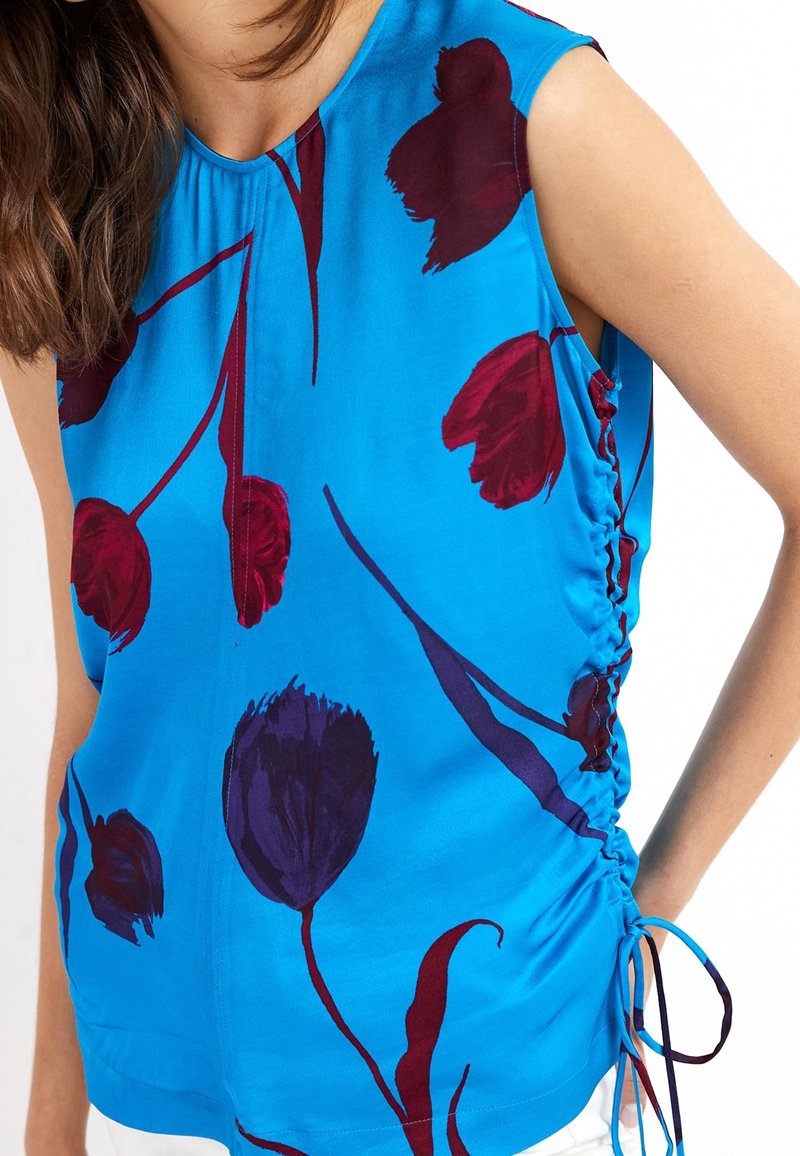 Blauw bloemen top met rode en donkerpaarse tulp patronen, korte mouwen en zijruche met verstelbare strikken. Glad stoffen textuur.