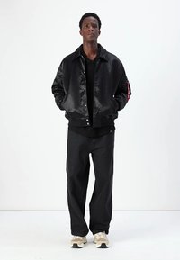 Casaco bomber preto com acabamento brilhante, padrão texturizado, fecho de pressão, bolsos laterais, combinado com calças pretas e sapatos beges.