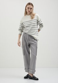Junge Frau, die einen creme- und grau gestreiften Pullover, graue Cargo-Hosen mit Bündchen und schwarze Slipper trägt, steht vor einem schlichten Hintergrund.