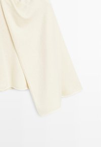 Haut à manches longues couleur crème avec un poignet doux et légèrement évasé, présenté drapé sur un fond blanc. Aucun motif ni ornement visible.