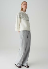 Maglione crema con un giogo grigio a motivi, caratterizzato da un colletto alto e polsini arrotolati. Abbinato a pantaloni larghi grigio chiaro e scarpe scure.