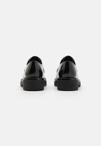 Paire de mocassins en cuir verni noir avec talons massifs, vue de dos sur un fond blanc uni.