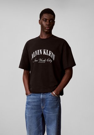 Ung mand med mørk hud iført en sort Calvin Klein New York City T-shirt og blå jeans, hænderne i lommerne, neutral baggrund.