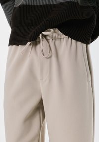 Bež elastične hlače s bočnim džepovima, strukom na uže i glatkom teksturom, uparen s tamno prugastim puloverom.