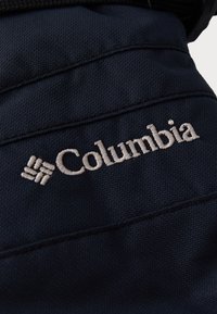 Mørkeblått stoff med en sydd Columbia-logo i lys beige. Teksturert overflate med synlige sømmer og detaljer.