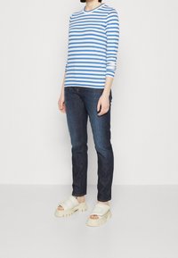 Camicia a maniche lunghe a righe blu e bianche in cotone, abbinata a jeans blu scuro e sandali a ciabatta cremosi con suole rigate.