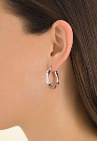 Orecchini a cerchio in argento con una forma irregolare e scultorea, caratterizzati da un piccolo dettaglio a perno. Superficie in metallo liscia e riflettente.