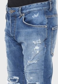 Jeans in denim blu strappati con un lavaggio sbiadito, caratterizzati da molteplici tasche, cuciture visibili e bordi sfrangiati per un aspetto usurato.