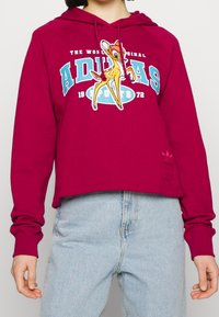 Person wearing a maroon hoodie with a cartoon deer and “ADIDAS SPORTS 1972” text, paired with light blue jeans.