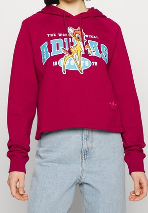 Persona che indossa un hoodie bordeaux con un cervo da cartone animato e la scritta "ADIDAS SPORTS 1972", abbinato a jeans azzurri chiari.