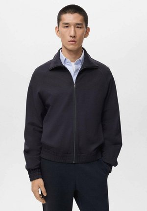 Dark blue zip-up jacket with a high collar and ribbed cuffs. Smooth fabric with a minimalistic design, worn over a light blue collared shirt.