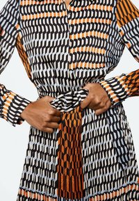 Des mains ajustant la ceinture d'une robe à manches longues avec un motif géométrique noir, blanc, orange et bleu clair.