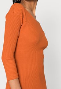 Top orange côtelé avec un décolleté en V et des manches trois-quarts, offrant une silhouette ajustée et une texture lisse.