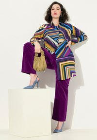 Multikleurige gestreepte blouse, paarse fluwelen broek, blauwe slingback hakken. Houdt een gele gestructureerde handtas vast, geposeerd op een witte sokkel.