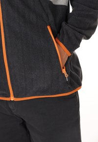 Fleecejakke i mørkegrå med orange lynlåse og manchetter. Har ribbet tekstur og sidelommer med lynlås.