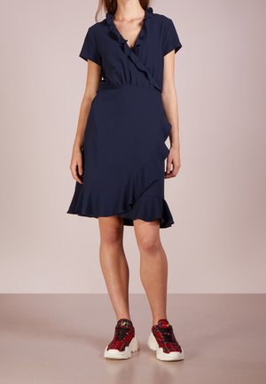 Mujer que lleva un vestido azul oscuro de manga corta con volantes, combinado con zapatillas gruesas rojas de cuadros, de pie frente a un fondo liso.