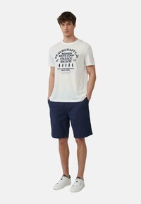 Witte katoenen T-shirt met marineblauwe grafische tekst, gecombineerd met marineblauwe shorts en witte sneakers. Het shirt heeft een strandthema print.
