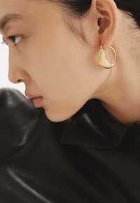 Pendiente de aro de oro con un diseño ondulado, superficie texturizada y un acabado pulido, llevado en la oreja de una persona sobre un fondo de cuero oscuro.