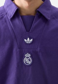 Lila tröja med V-ringad design. Har broderade logotyper för adidas och Real Madrid i vitt. Tillverkad av slitstarkt tyg med en texturerad yta.
