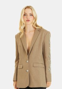 Blazer marrón entallado con un escote en V profundo, dos botones y patrones de hojas adornadas en las mangas, hecho de una tela texturizada.