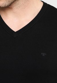 Svart stickad tröja med v-ringad halsringning och ribbad textur, med en liten tonad logo på bröstet, som visar en kroppsnära design.