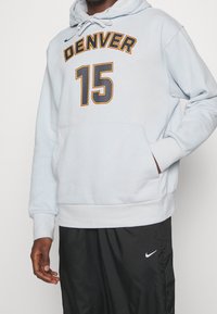 Hombre vestido con una sudadera ligera gris de Denver con el número 15 y pantalones negros de Nike, de pie con una mano en el bolsillo de la sudadera.
