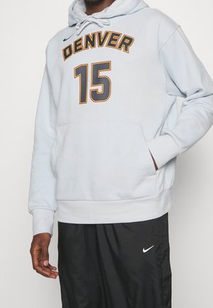 Uomo che indossa una felpa grigia chiara di Denver con il numero 15 e pantaloni neri Nike, in piedi con una mano nella tasca della felpa.