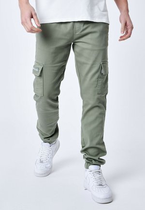 Pantalones cargo en verde oliva, hechos de tela suave, con un ajuste entallado, bolsillos laterales y puños elásticos. Llevados con zapatillas blancas.
